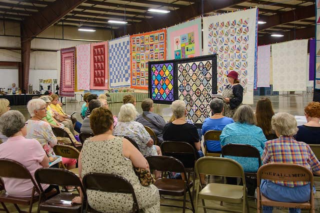 Ladies sitting in chairs in front of a row of quilts. Copyright: HolmanPhotos.com