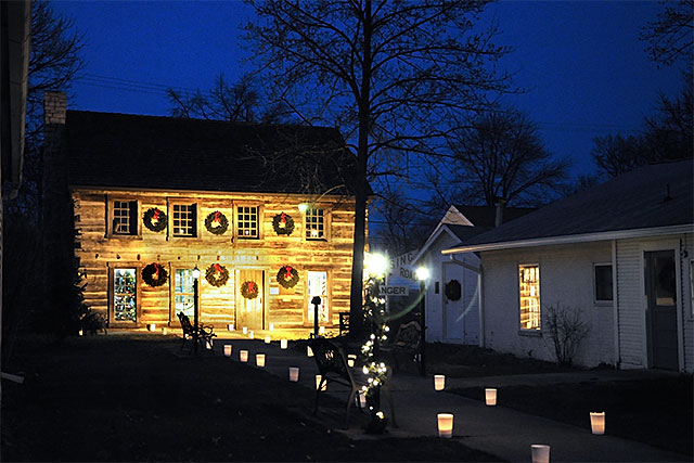 Photo: Log Cabin at dusk with Christmas wreaths on the outside and luminarias on the sidewalk in front of it.